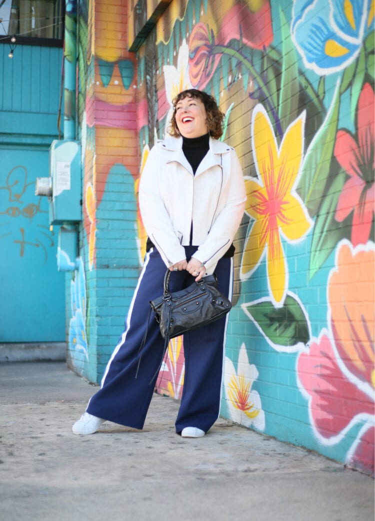 Alison in a white leather jacket, black turtleneck, and navy wide leg pants holding a black handbag in front of her.