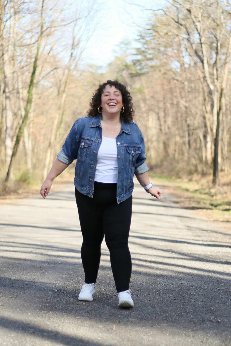 Alison of Wardrobe Oxygen laughing and dancing on a gravel road while wearing black leggings with a denim jacket and white Allbirds sneakers