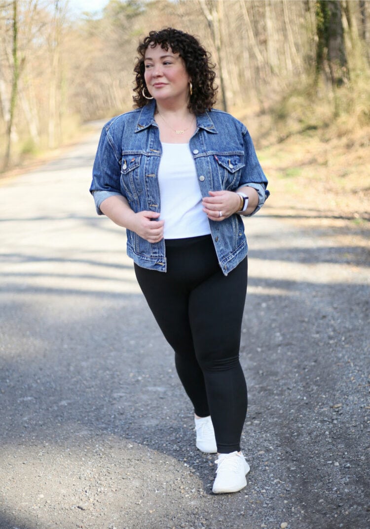 Alison of Wardrobe Oxygen laughing and dancing on a gravel road while wearing black leggings with a denim jacket and white Allbirds sneakers