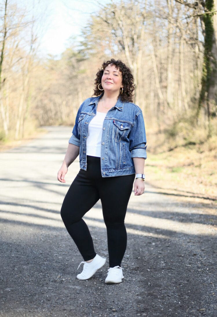 Alison of Wardrobe Oxygen in black leggings, a white tank, and denim jacket grinning while standing on a gravel road through a wood