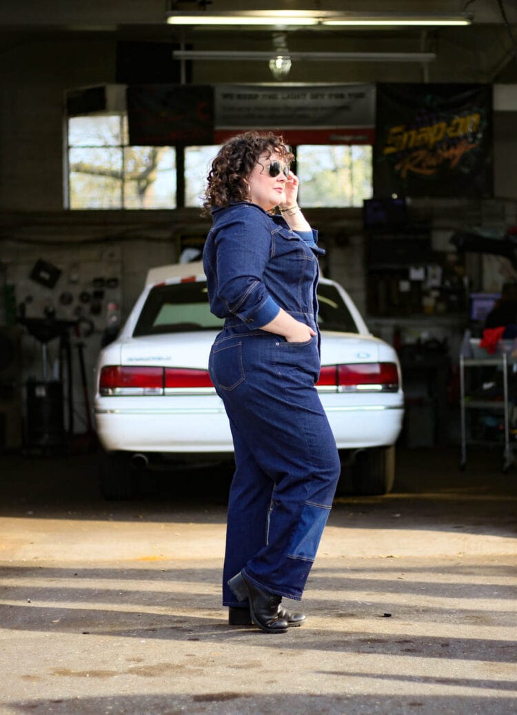 Alison in a dark denim pair of coveralls or a boilersuit standing in front of an auto body shop