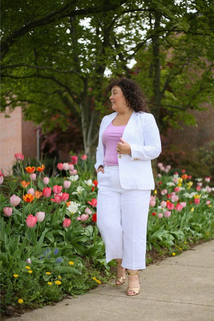 Talbots Linen Pantsuit: White with Lavender 1 Alison in a Talbots linen blazer and cropped pants with a lavender knit shell underneath, walking along a sidewalk flanked by tulips