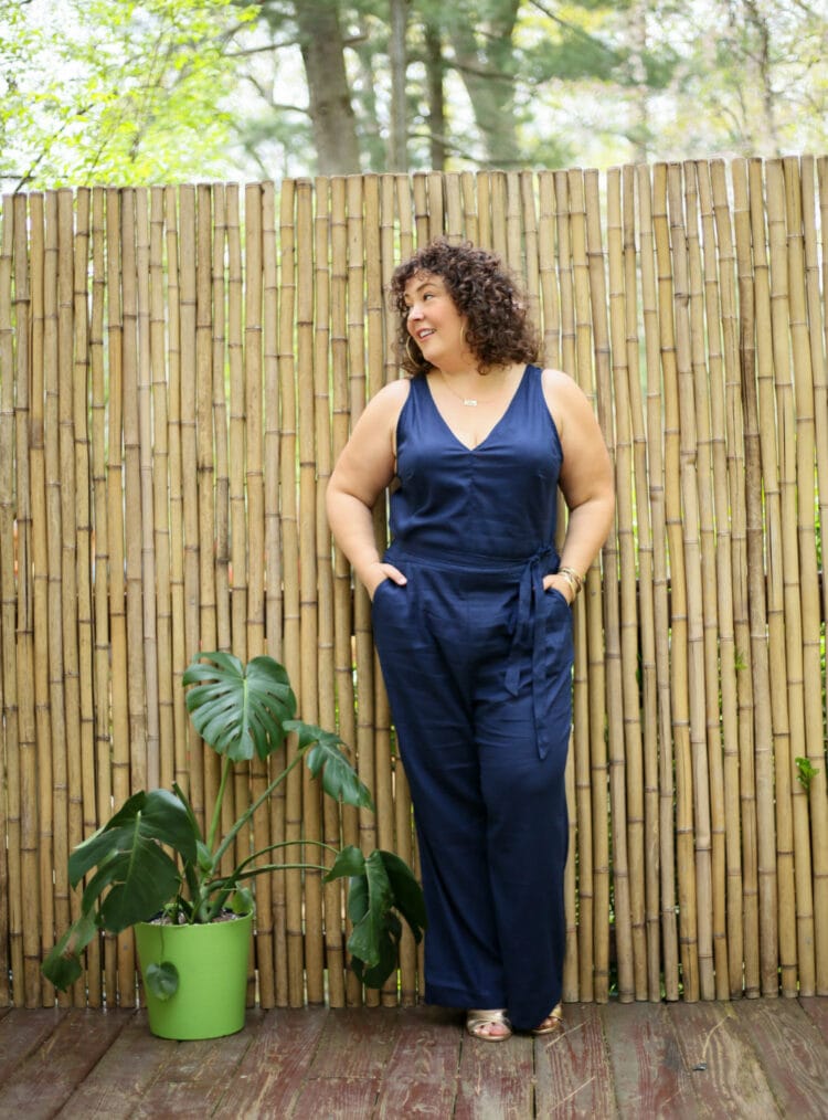 Alison wearing a navy sleeveless linen jumpsuit from Universal Standard that has a v-neck and a self belt. SHe paired it with gold Margaux heeled sandals