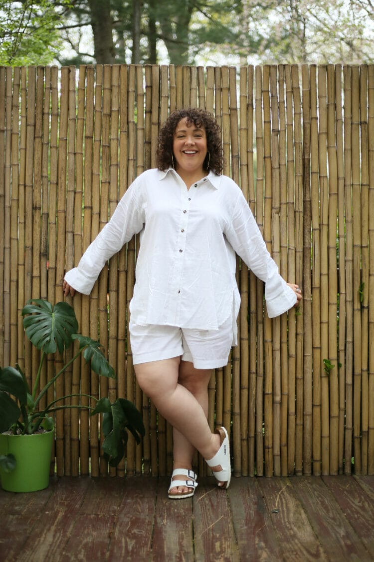 Universal Standard linen long-sleeved shirt and loose above-knee shorts on Alison. She is standing in front of a wall of bamboo with her legs crossed and smiling at the camera.