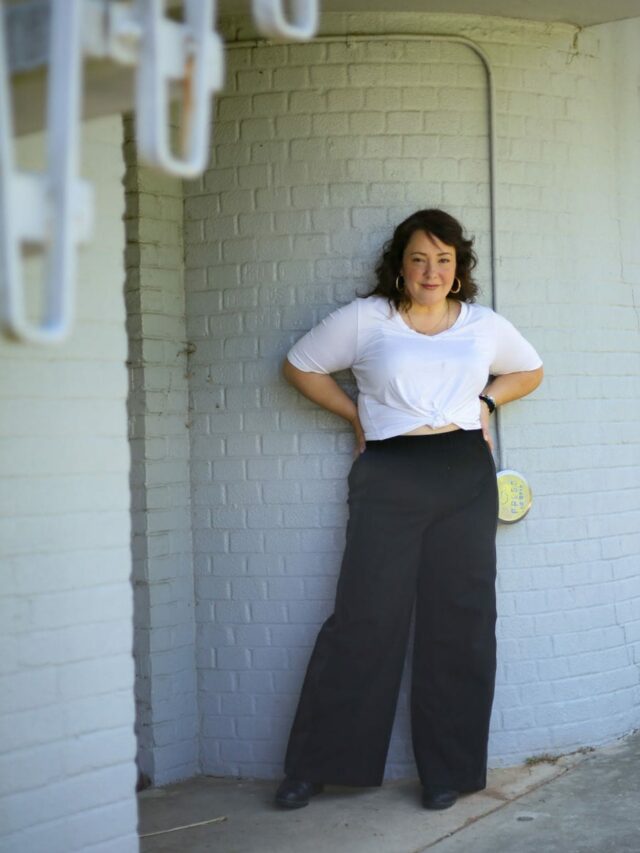 Alison in a white v-neck top tied at the waist with black wide-leg pants from Universal Standard. She is standing against a gray brick wall with her hands on her hips.