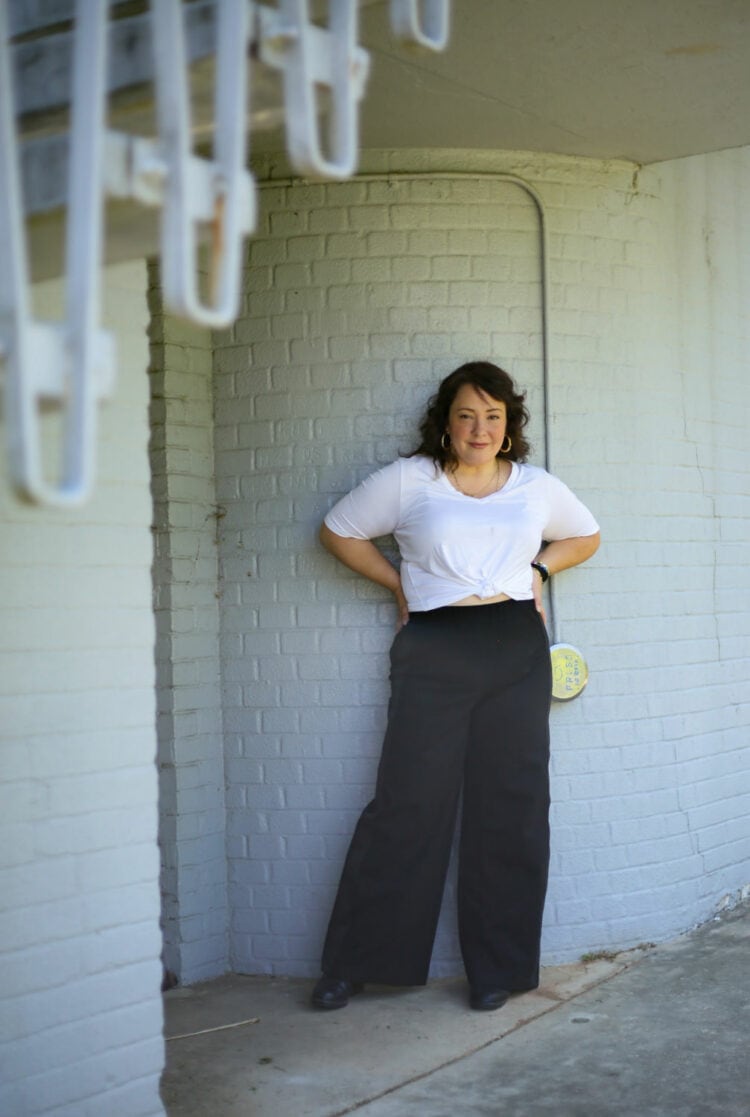 Alison in a white v-neck top tied at the waist with black wide-leg pants from Universal Standard. She is standing against a gray brick wall with her hands on her hips.