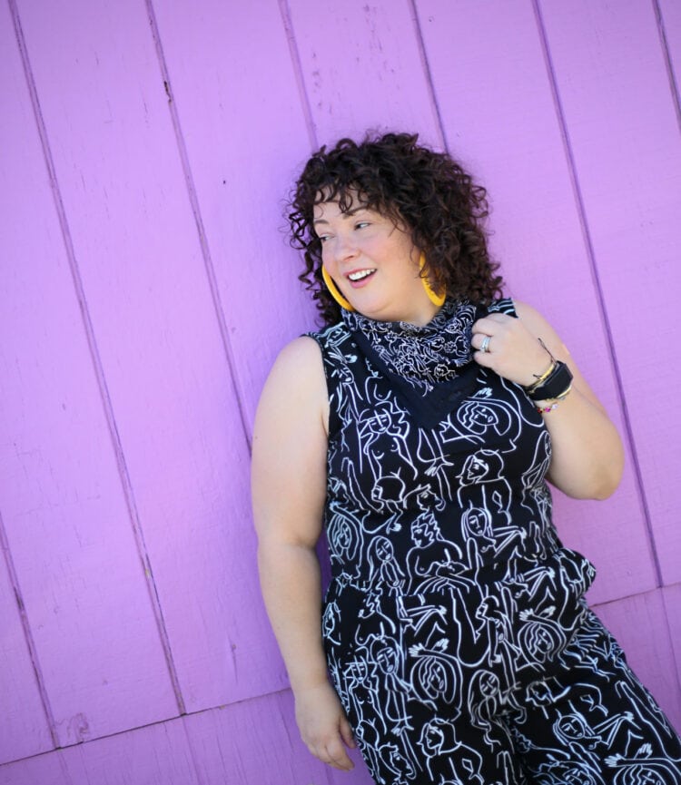 Woman with curly brown hair and biog yellow hoop earrings leaning against a purple wood wall. She is wearing a black and white graphic print top, pants, and bandana from Dazey LA