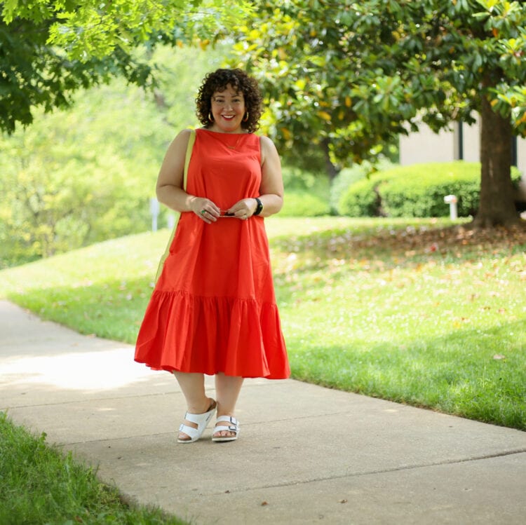 Alison wearing the Christopher John Rogers for Target Orange Shift Dress with white Birkenstocks and a lemon yellow leather camera bag from Talbots. She is holding a pair of Ray-Ban aviators in her hands and smiling at the camera as she walks down a sidewalk on a tree-lined path.