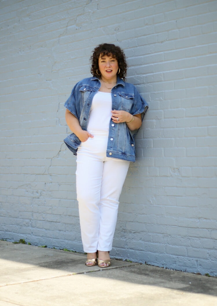 Alison of Wardrobe Oxygen wearing a denim ruana from Chico's with white straight jeans and a white tank and gold Margaux Uptown Sandals. She is standing against a pale gray brick wall, one hand in a pocket, looking at the camera.