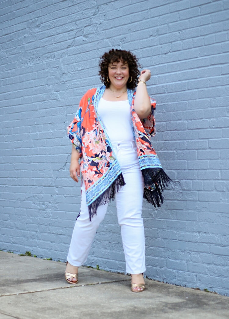 Alison in an orange printed linen summer ruana from Chico's with white jeans and a white tank. She is laughing at looking at the camera, her left hand in her hair.