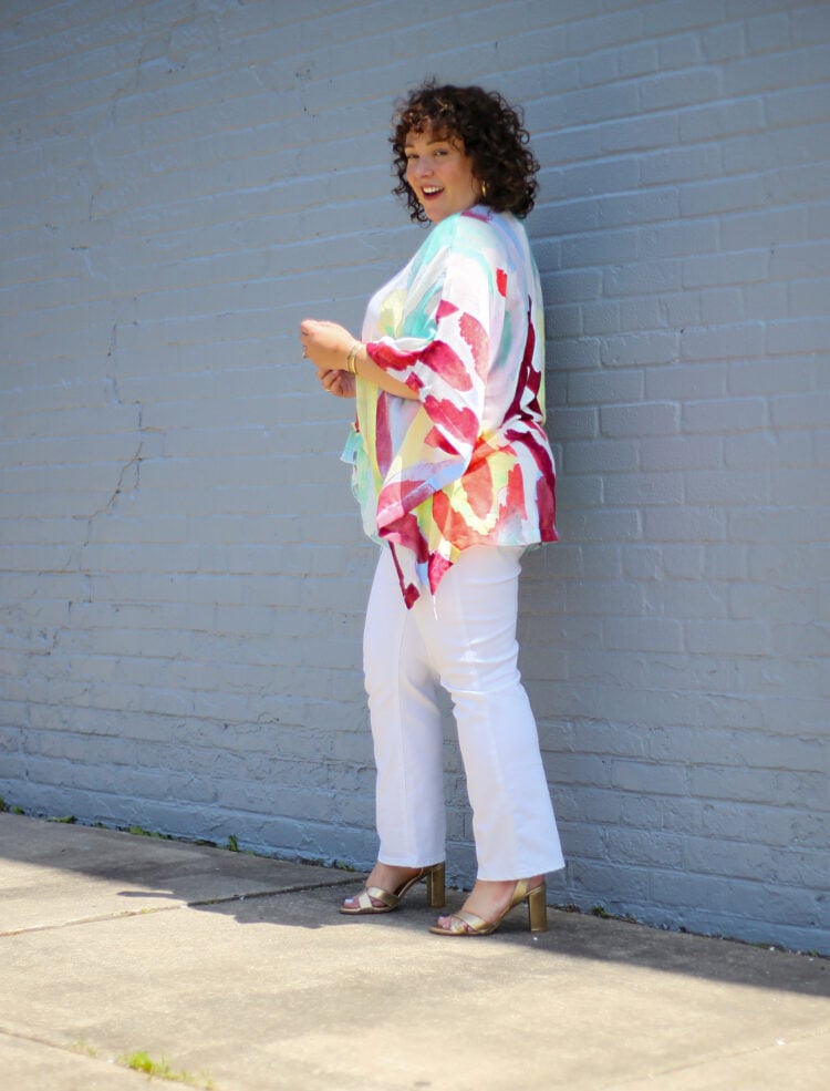 Alison of Wardrobe Oxygen in a lightweight summer ruana from Chico's that is white with a watercolor brush stroke design in reds and greens and yellows. She is wearing it with white jeans and is walking away from the camera but looking back and smiling