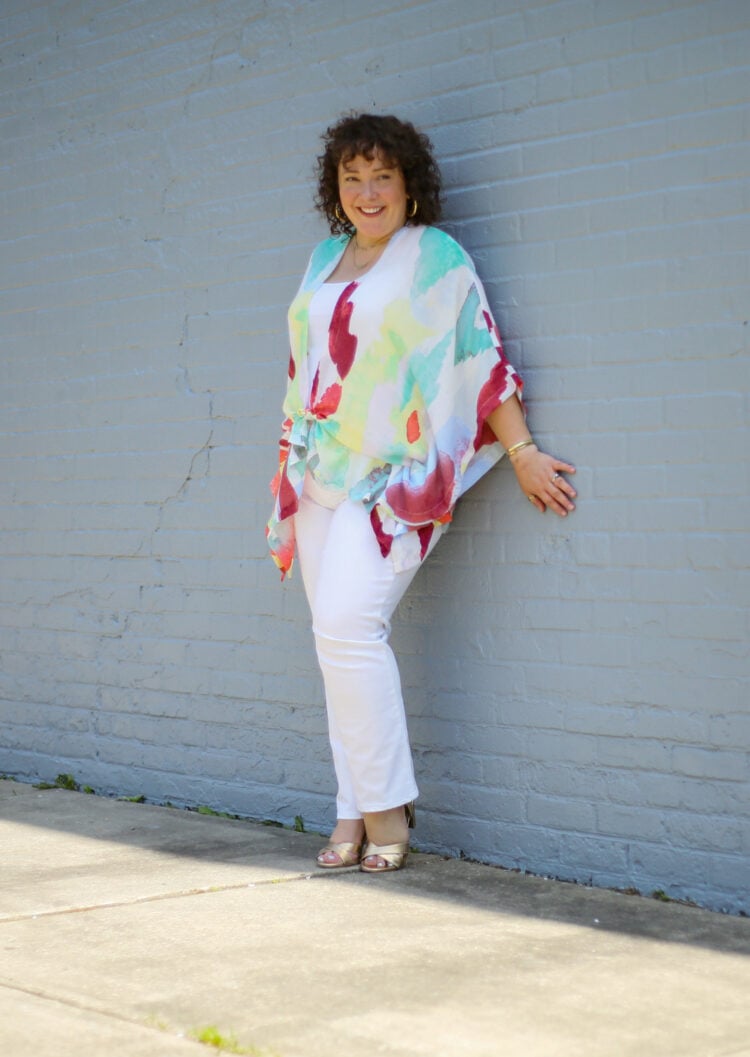 Alison of Wardrobe Oxygen styling a summer ruana from Chico's with white jeans. She is leaning against a gray wall and looking to the left