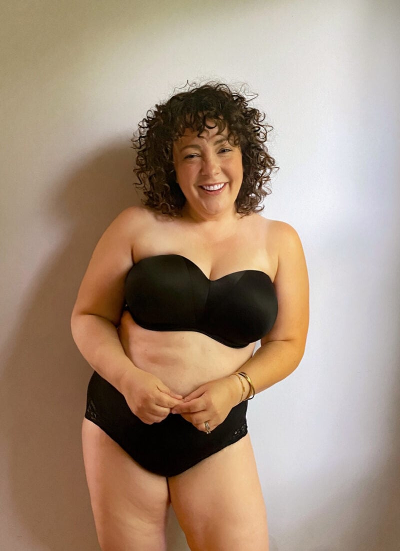 Alison wearing a black Soma Stunning Starlet strapless bra with black briefs. She is standing against a wall and smiling at the camera
