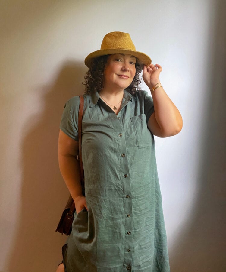 Alison wearing a sage green linen shirtdress with cap sleeves. She is wearing a straw hat and touching the brim, a brown handbag on her shoulder