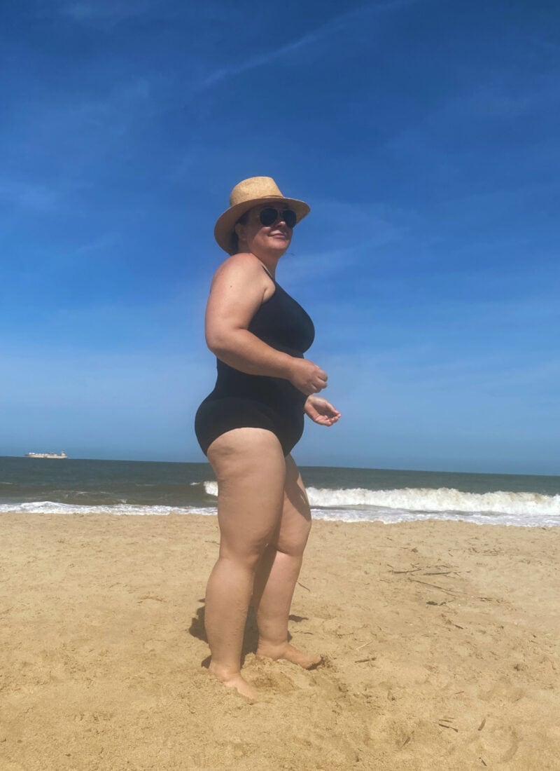 Is this the best swimsuit for women over 40? Alison of Wardrobe Oxygen models the Lands' End Tugless tank in black. She is standing on the beach facing to the right and is smiling at the camera.
