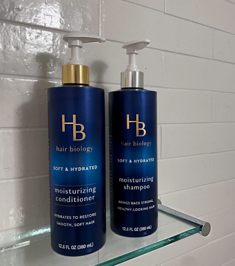 Bottles of Hair Biology Moisturizing shampoo and conditioner on a glass shelf inside a white tiled shower.