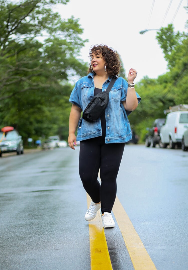 Alison standing in the middle of a street. She is wearing a hip length sleeveless denim jacket and black leggings. She has on white Adidas Superstar sneakers and has a black belt bag across her body.