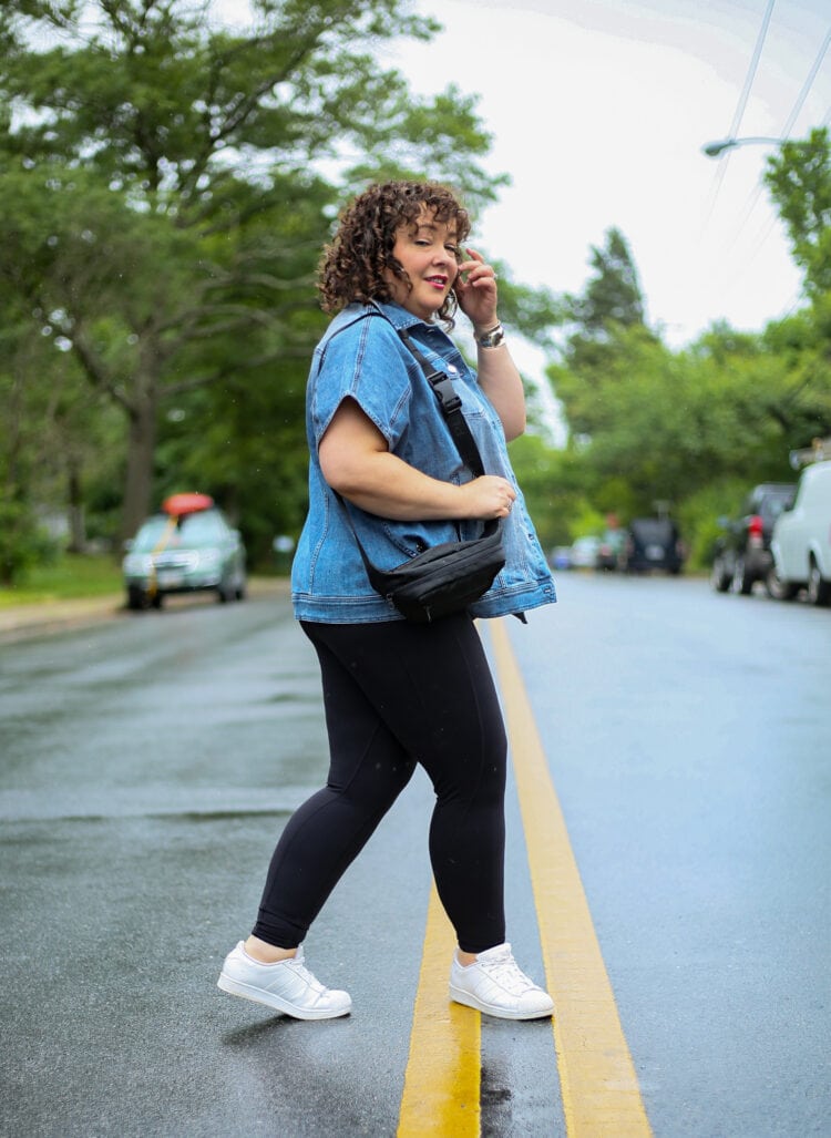 Alison in the Universal Standard Next to Nothing Bodysuit styled with a denim ruana and white sneakers. She is walking across a street looking at the camera.