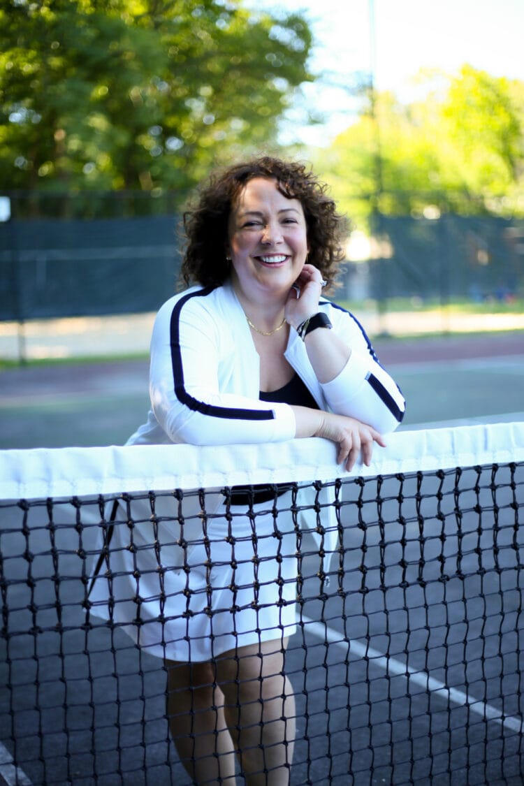 Alison in a white knit bomber jacket and matching skort from Talbots. She is leaning against a tennis net smiling at the camera