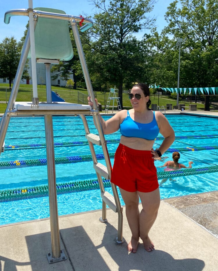 Alison in the Universal Standard Sunny Swim Shorts in red with a turquoise bikini top. She is standing in front of a public swimming pool wearing Ray-Ban aviators.