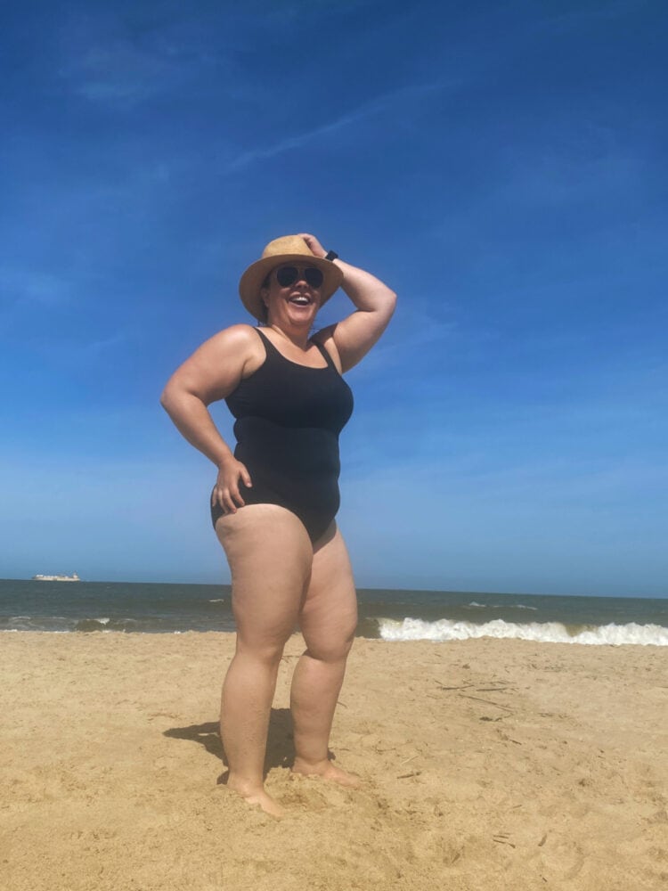 Alison in the Lands' End Tugless Tank swimsuit in black. She is standing on the beach with her hand on her head laughing to the camera.