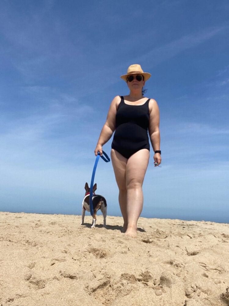 IMO the Best Swimsuit for Women Over 40 1 Wardrobe Oxygen wearing the Lands' End Tugless Tank in black with a straw fedora and aviator sunglasses. She is standing on the beach with her dog on a leash.