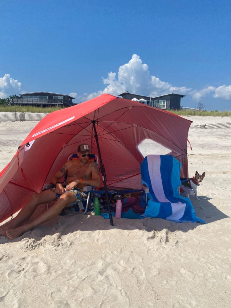 The beach of Davis Park Fire Island with a red oversized umbrella shade shelter. Under the shelter are two beach chairs. One has a man in it, the other has a small dog