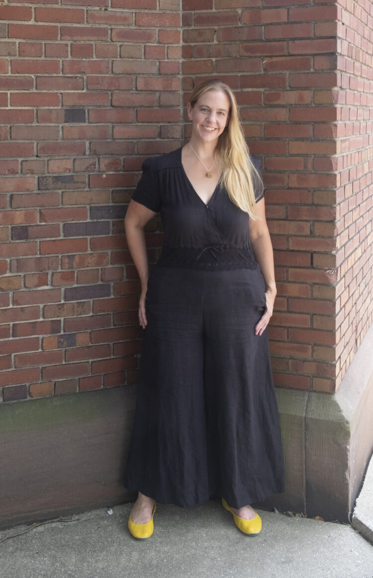 woman with long blonde hair wearing a black jumpsuit and yellow leather flats standing in front of a brick wall