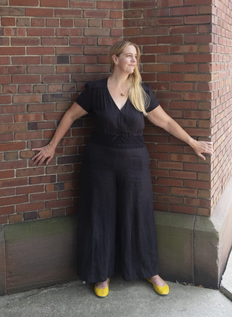 woman with long blonde hair wearing a black jumpsuit and yellow leather flats standing in front of a brick wall