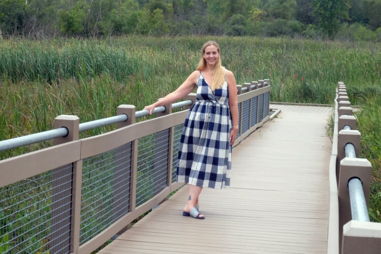 Woman with long blonde hair wearing a gingham printed sundress from Christy Dawn, standing o a bridge next to a field.