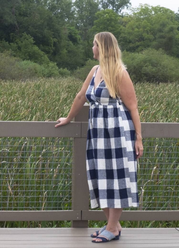 Woman with long blonde hair wearing a gingham printed sundress, standing o a bridge next to a field.