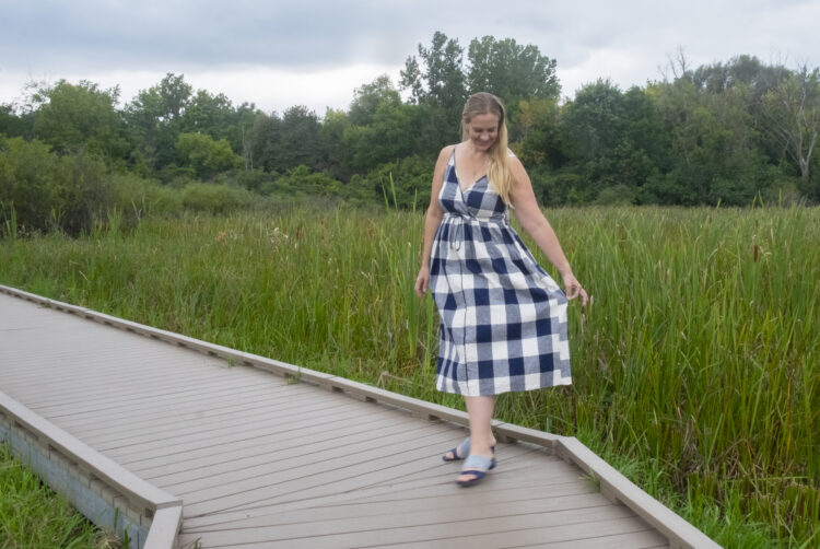Woman with long blonde hair wearing a gingham printed sundress from Christy Dawn, standing o a bridge next to a field.