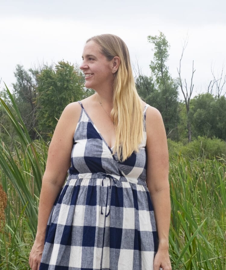 Woman with long blonde hair wearing a gingham printed sundress from Christy Dawn, standing o a bridge next to a field.