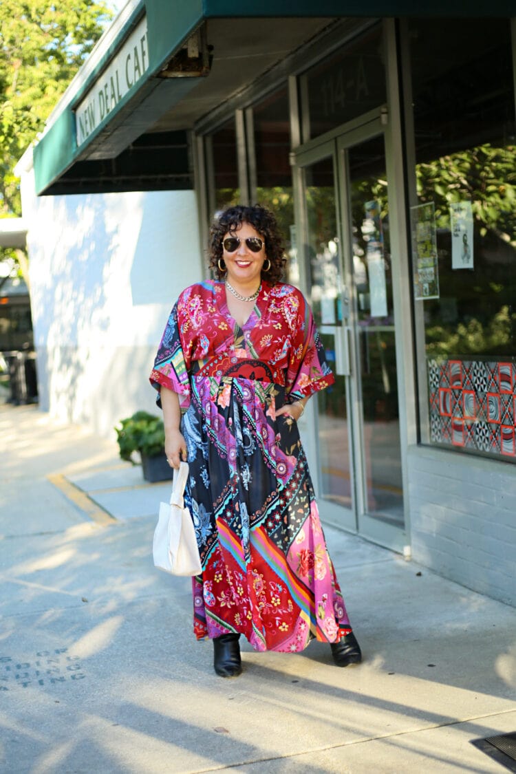 Woman in a red printed maxi dress from eShakti, paired with black heeled knee high boots and a cream leather pouch handbag. She is wearing sunglasses and smiling. She is standing on a sidewalk outside a cafe.