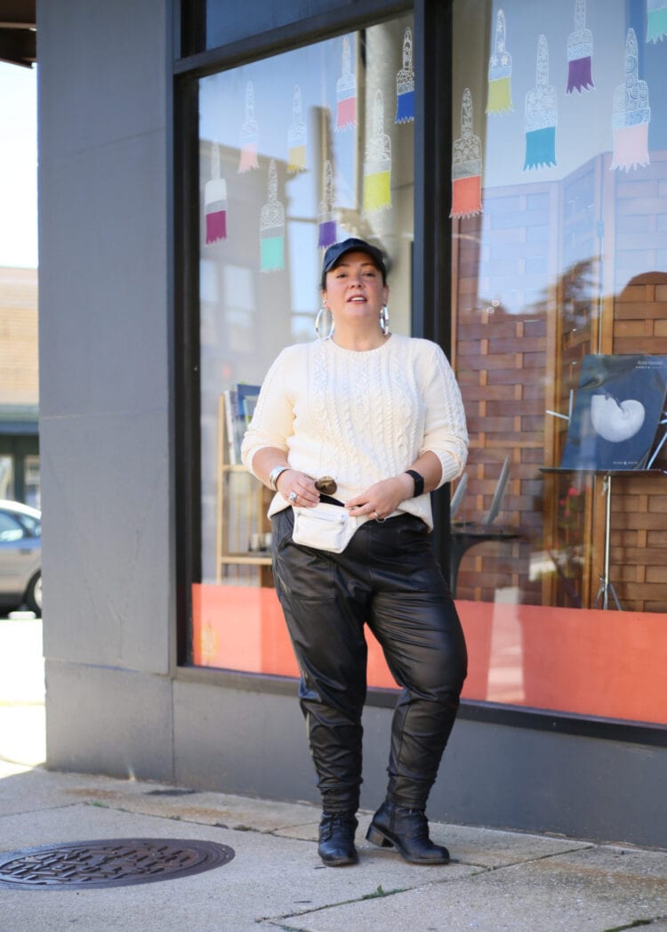 woman in a black faux leather baseball cap, oversized silver hoops, cream cableknit crewneck sweater, black faux leather joggers, and black lace up ankle boots