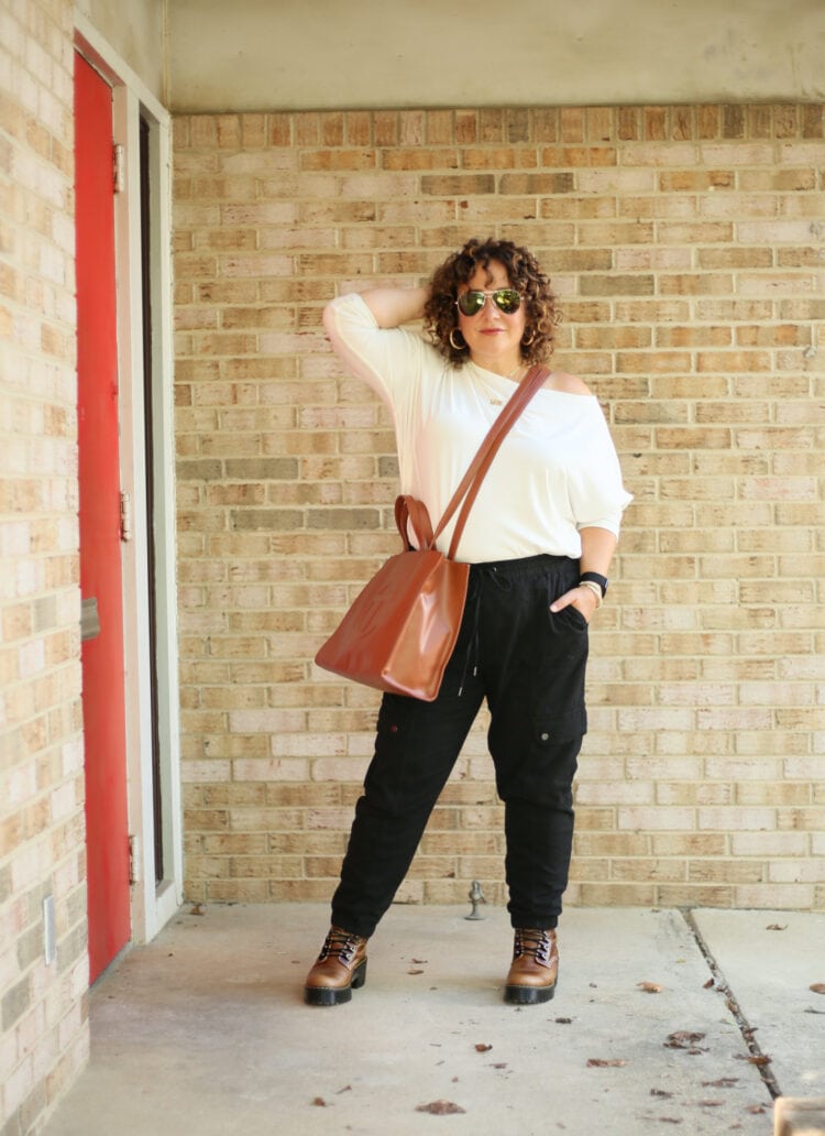 ALLSAINTS Rita Off the Shoulder Top with a Medium TELFAR Bag 1 Wardrobe Oxygen in a white ALLSAINTS Rita off the shoulder long sleeved top with black jogger inspired black cargo pants from Banana Republic and a tan TELFAR medium tote worn as a crossbody. She is standing in front of a brick wall wearing sunglasses and facing the camera, one hand in her hair.