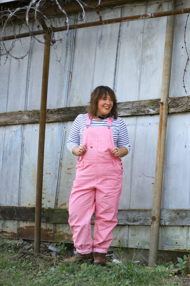 Alison of Wardrobe Oxygen in pink Dickies overalls and a black and white striped long sleeved tee. She is standing in front of a metal storage unit with her hands in her pockets smiling away from the camera