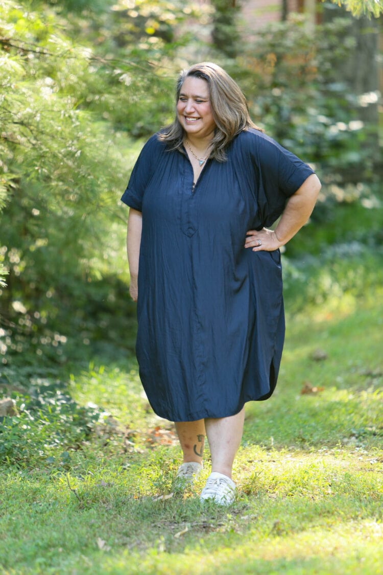 Carrie wearing a navy ever by X dress unbuttoned and loose. She is standing in a field with her left hand on her hip and she is laughing.