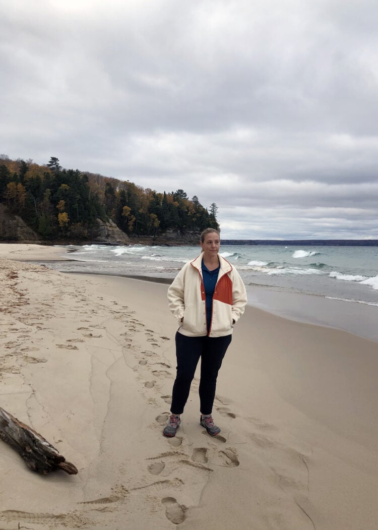 Lissa wearing the alder apparel go far fleece with a teal shirt and black pants. She is standing on a beach with trees and the shore behind her.
