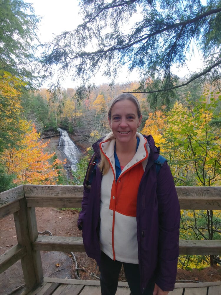 Lissa wearing the alder apparel go far fleece while hiking. Over the fleece she is wearing a purple raincoat and a teal hiking backpack. She is standing in front of a waterfall smiling for the camera.