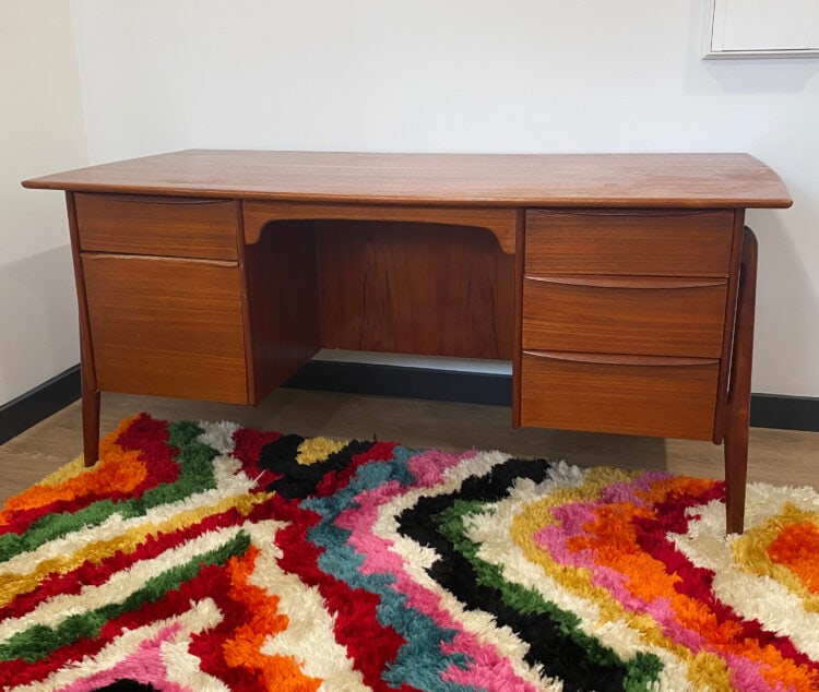 Svend Åge Madsen teak executive desk with five drawers in the corner of Alison's office. It is sitting on a multicolored shag carpet against a plain white wall.