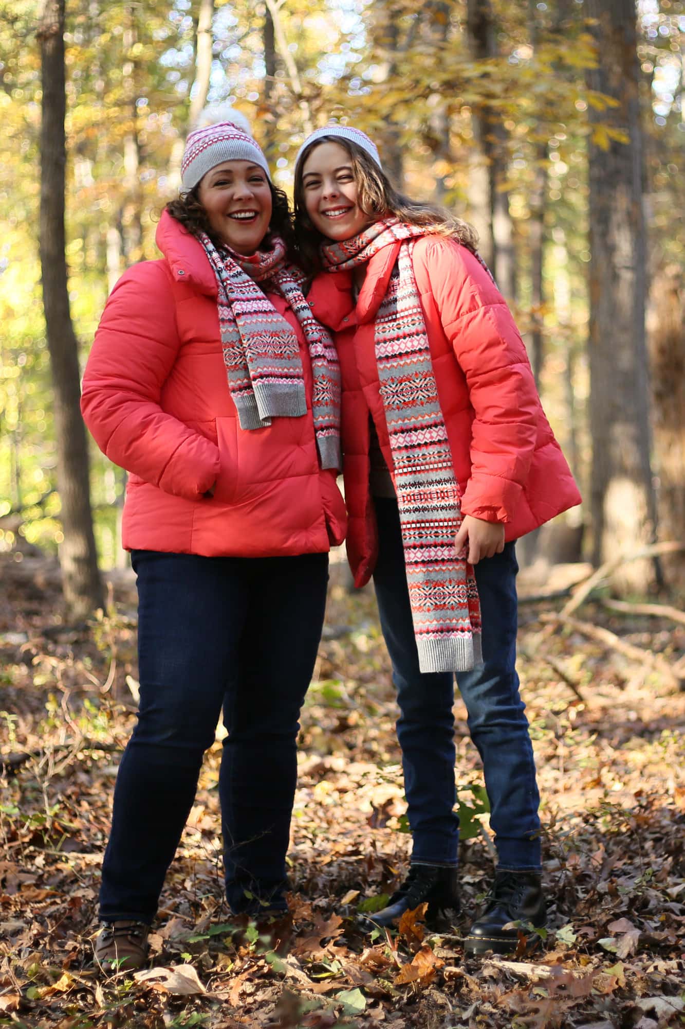 Alison and her Daughter hugging in the woods, smiling at the camera. The sun is shining on her daughter's shoulder. They are wearing buttoned up coral colored hip length puffer coats and Fair Isle print scarves wrapped around their necks twice. They are also wearing Fair Isle print beanie caps with a gray cuff and white furry pom pom on top.