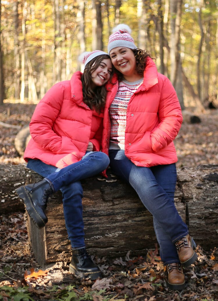Alison and her 12-year-old daughter sitting on a log in the woods. They both have their legs crossed and are smiling, Alison's daughter has her head on Alison's shoulder. They are wearing coral colored puffer jackets open over Fair Isle print sweaters and are wearing matching beanie caps.