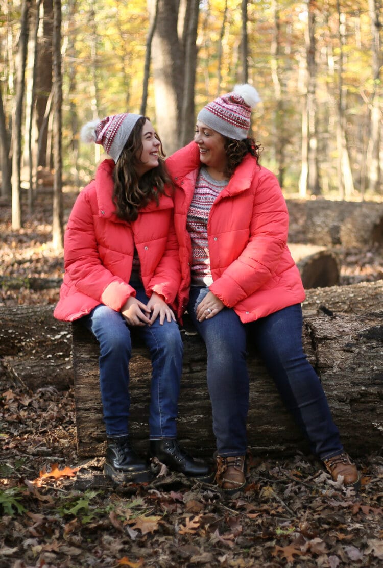 Alison and her daughter in the woods sitting on a fallen log. They are both wearing coral colored short puffer jackets with coral and gray Fair Isle printed crewneck sweaters, scarves, and beanies all from Talbots. They are wearing jeans and Doc Marten boots and are smiling at one another.