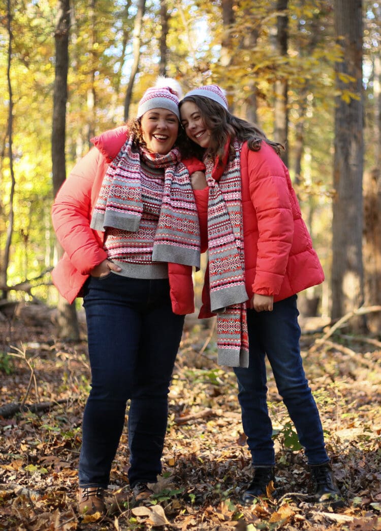 Alison and her daughter are standing in the woods and sun is dappled on their faces. They have their arms around one another and are smiling at the camera, their temples touching. They are wearing matching outfits od dark slim-fitting jeans, coral-colored puffer coats that hit mid-hip, and Fair Isle printed sweaters, long scarves, and knit caps of the same coral, white, and gray color. 