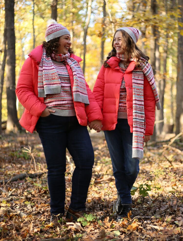 Alison and her daughter walking in the woods, holding hands and smiling at one another. They are both wearing coral colored short puffer jackets with tall collars and dark jeans. They are also wearing coral and gray Fair Isle printed sweaters, scarves, and beanies. All the clothing is from Talbots.