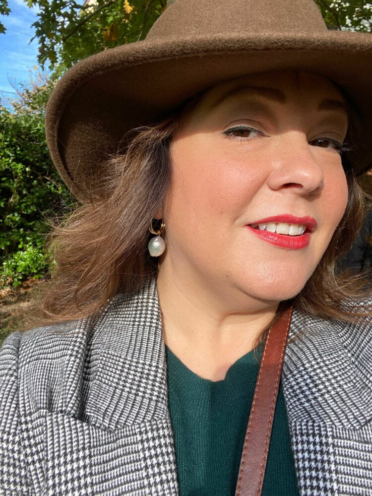 Alison looking at the camera wearing a brown bolero hat and gray plaid blazer. She is wearing red lipstick and gold hoop earrings from The Freshwater Pearl Company and each hoop has a freshwater pearl drop hanging from it.