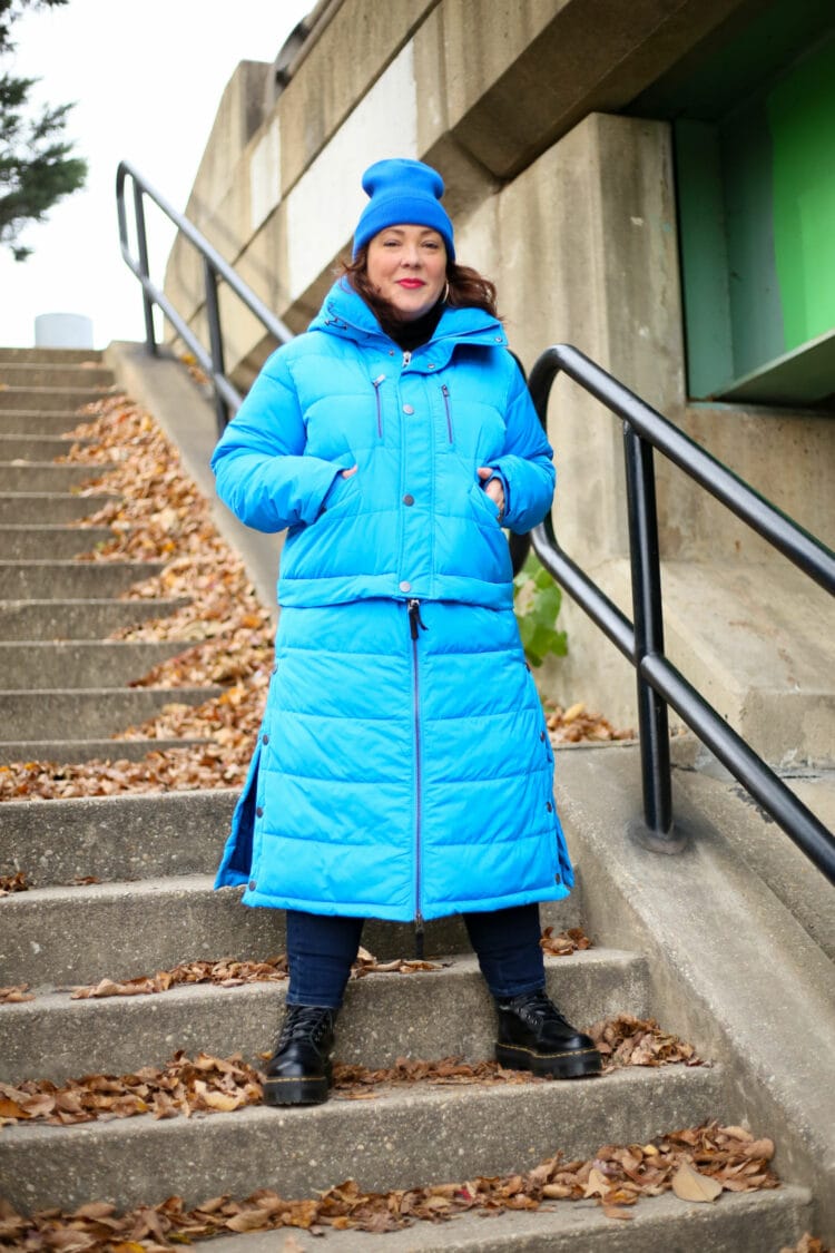 Alison wearing the full length version of the Universal Standard 4-in-1 Hybrid Puffer in electric blue. She has her hands in the pockets, she is standing wide on an outdoor concrete stairwell, looking off into the distance. Her hair is covered with a cobalt blue knit beanie also from Universal Standard.