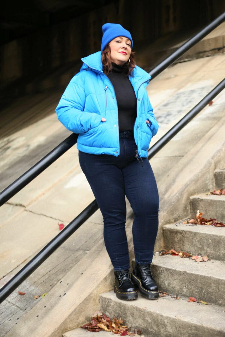 Alison leaning against a black railing on a concrete stairwell. She is wearing the electric blue puffer without the skirt attached. It is unzipped over a black turtleneck and dark rinse skinny jeans. She is smirking at the camera, wearing a cobalt blue knit beanie on her head and black Doc Marten boots on her feet.