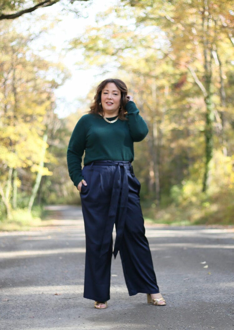 Alison in the green sweater and navy wide leg trousers walking towards the camera. She is tucking her shoulder length brown hair behind one ear.